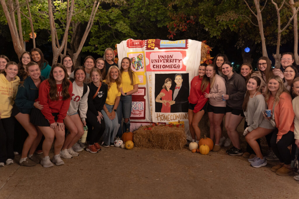 A group of young women pose in front of a decorated Chi Omega banner at night, surrounded by pumpkins and hay bales, with trees in the background. The scene appears to be part of a university homecoming event.