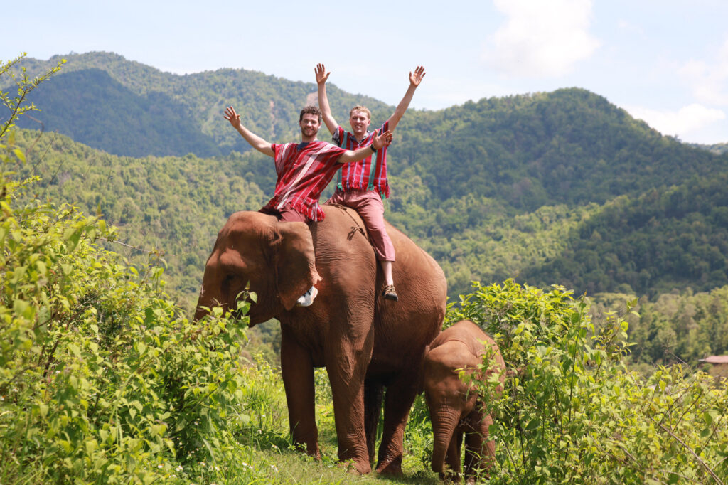 Two people wearing red-striped clothing sit on an adult elephant with their arms raised. A baby elephant stands nearby. They are outdoors in a green, hilly landscape under a partly cloudy sky.