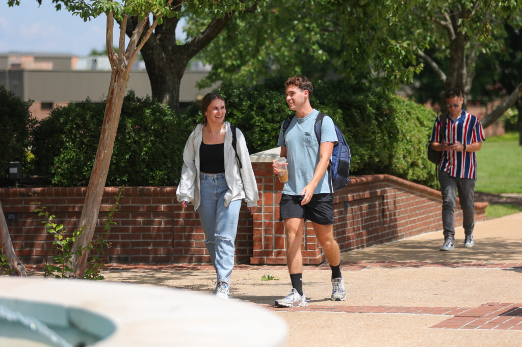 Two young adults walk on a campus path, talking and smiling. One holds a drink and wears a backpack. Trees and a brick wall line the path. Another person walks behind them.