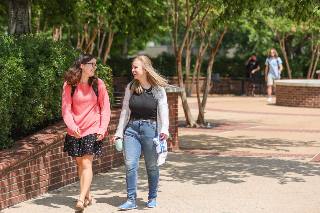 Two young women walk and talk on a sunny campus path, one carrying a backpack and the other holding a drink and snack. Trees and brick walls line the walkway, with more people in the background.