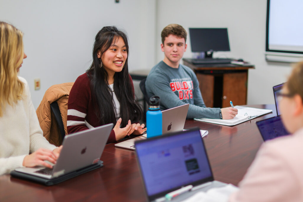 Four people are seated around a table with laptops and notebooks; one woman in a maroon sweater is speaking, while the others listen and take notes. A monitor and whiteboard are visible in the background.