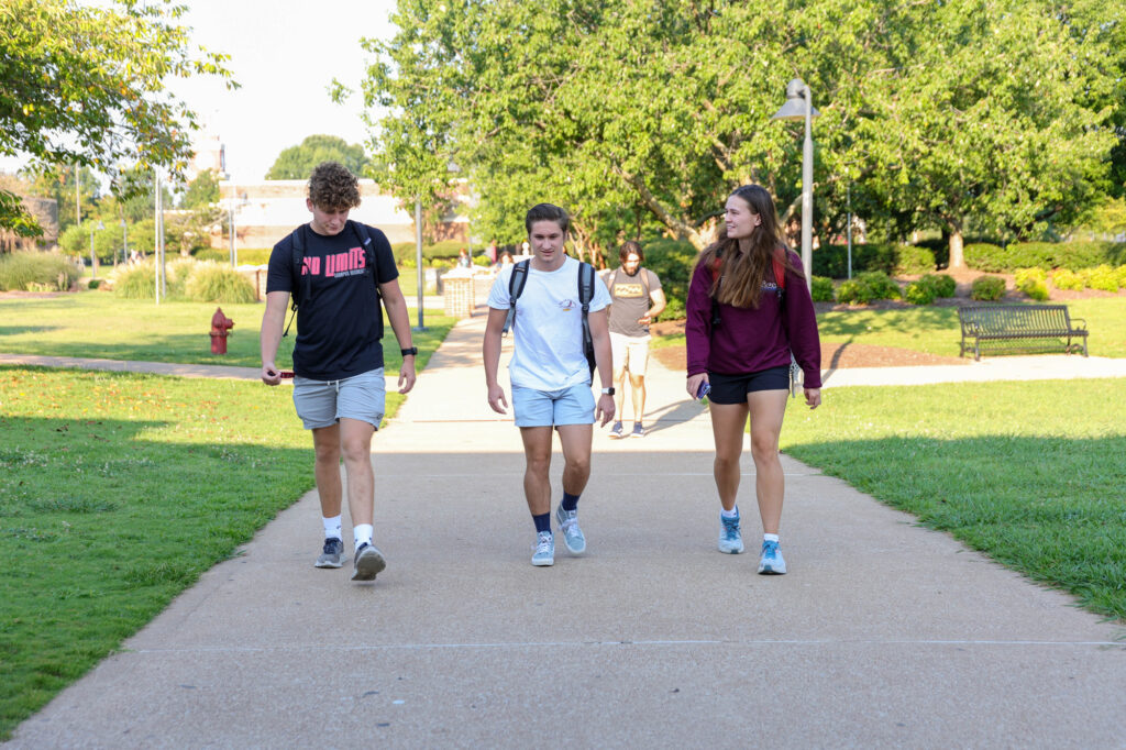 Three young adults walk on a paved path in a park-like setting with green grass and trees. Two men and one woman are in the foreground, and other people are visible in the background.