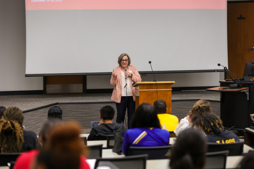 A woman stands at a podium speaking to an audience in a lecture hall. Several people are seated facing her. A large screen is displayed behind her.