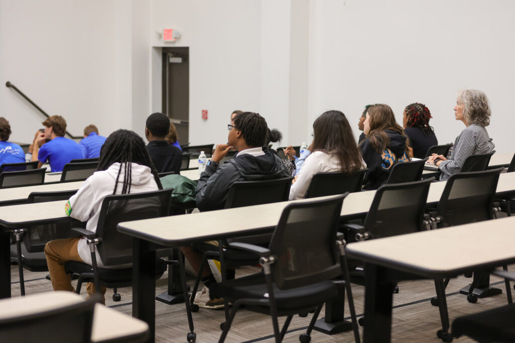 Students and one adult sit in rows of desks in a classroom or lecture hall, facing forward and listening. The room is brightly lit with empty desks in the foreground.