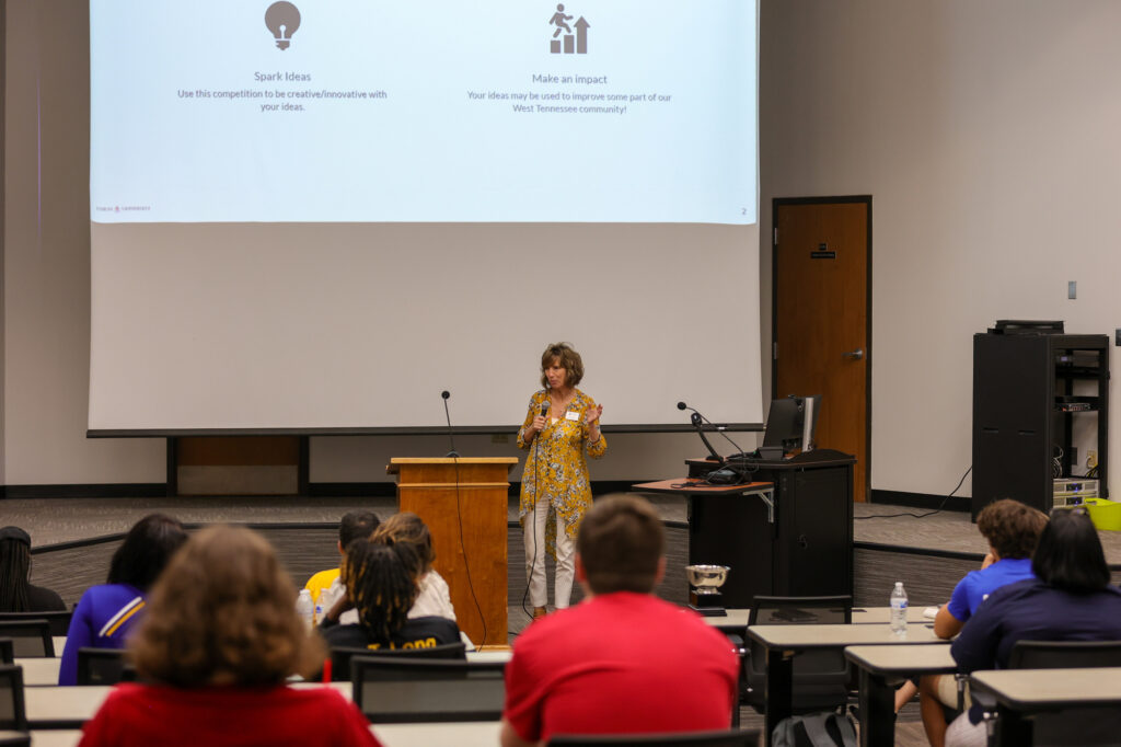A woman stands at a podium speaking to an audience in a lecture hall. A presentation slide behind her displays headings Spark Ideas and Make an Impact. Several people are seated facing the speaker.