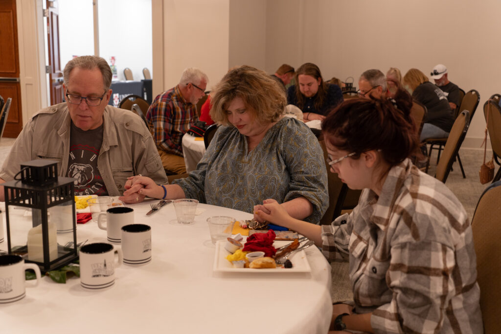 Three people sitting at a round table are holding hands with heads bowed, possibly in prayer, before a meal. Plates of food, mugs, and a lantern are on the table. Other people are seated at tables in the background.