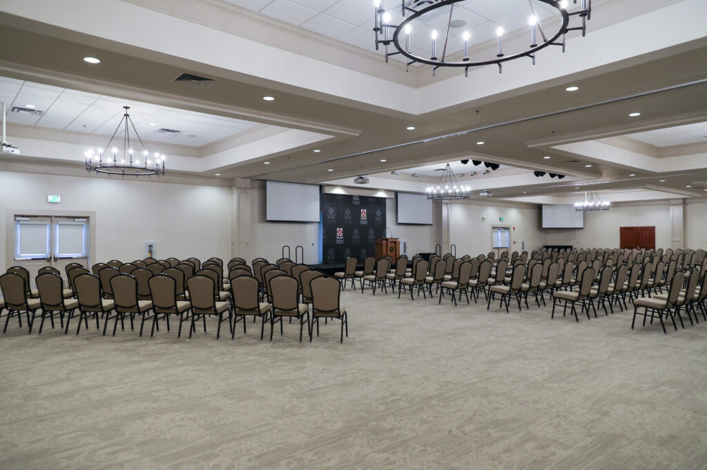 A large, empty conference room with rows of beige chairs facing a stage with a podium and black backdrop. Chandeliers hang from the ceiling, and multiple doors and windows are visible along the walls.