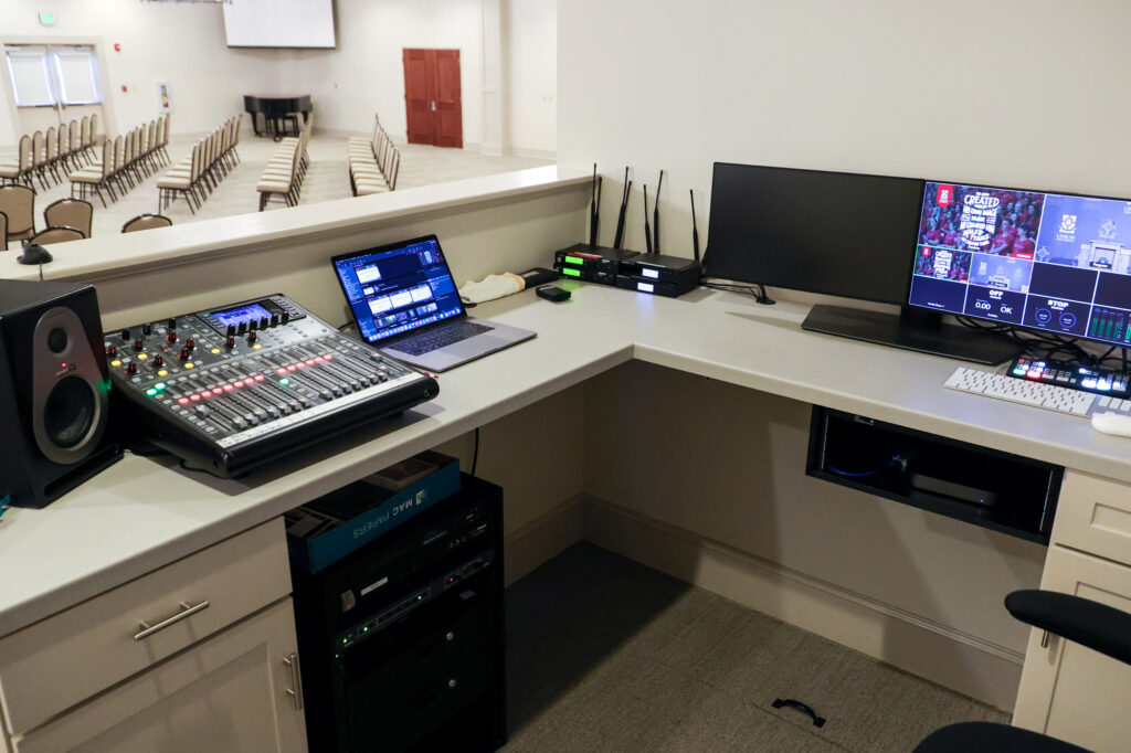 An AV control booth with a mixer, speaker, rack equipment, a laptop, a monitor displaying video feeds, routers, and a keyboard. Rows of empty chairs and a stage are visible in the background.