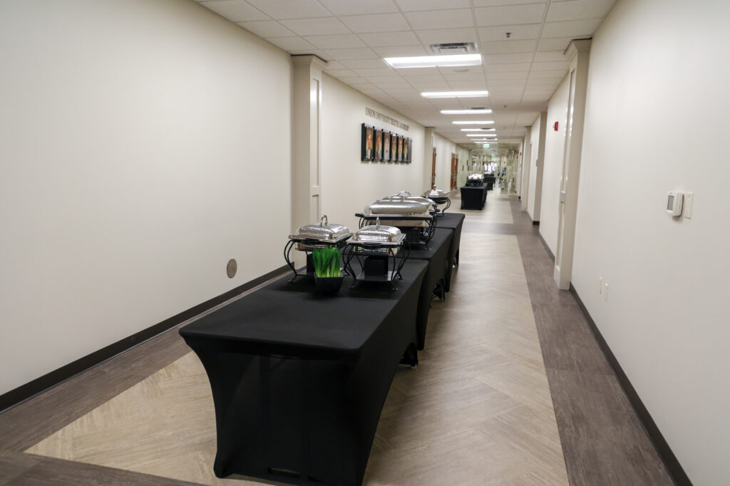 Two long black tables with covered chafing dishes and a small container of green utensils are set up in a hallway with beige walls and brown flooring. Framed photos hang on the left wall.