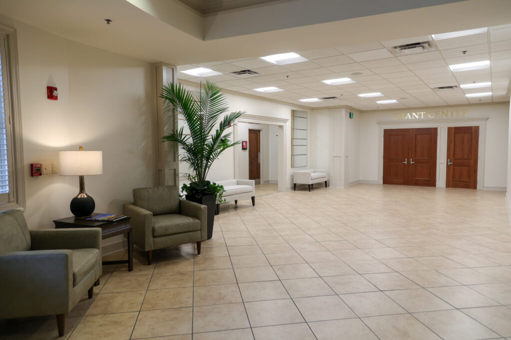 Spacious lobby with tiled floor, two green chairs and a lamp on the left, a potted plant, white benches along the walls, and double wooden doors labeled GIANT CENTER at the back.