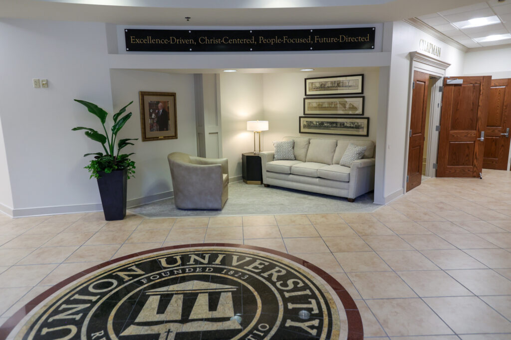 A university lobby features a large Union University seal on the floor, a small seating area with a couch, chair, lamp, wall art, potted plant, and a sign reading, “Excellence-Driven, Christ-Centered, People-Focused, Future-Directed.”.