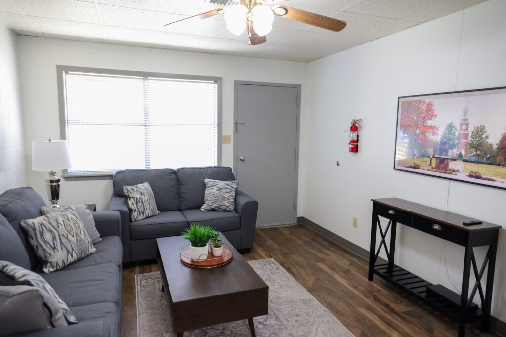 A small living room with two gray sofas, a wooden coffee table, a floor lamp, a wall-mounted TV, a console table, and a potted plant. Sunlight enters through a window next to a closed gray door.