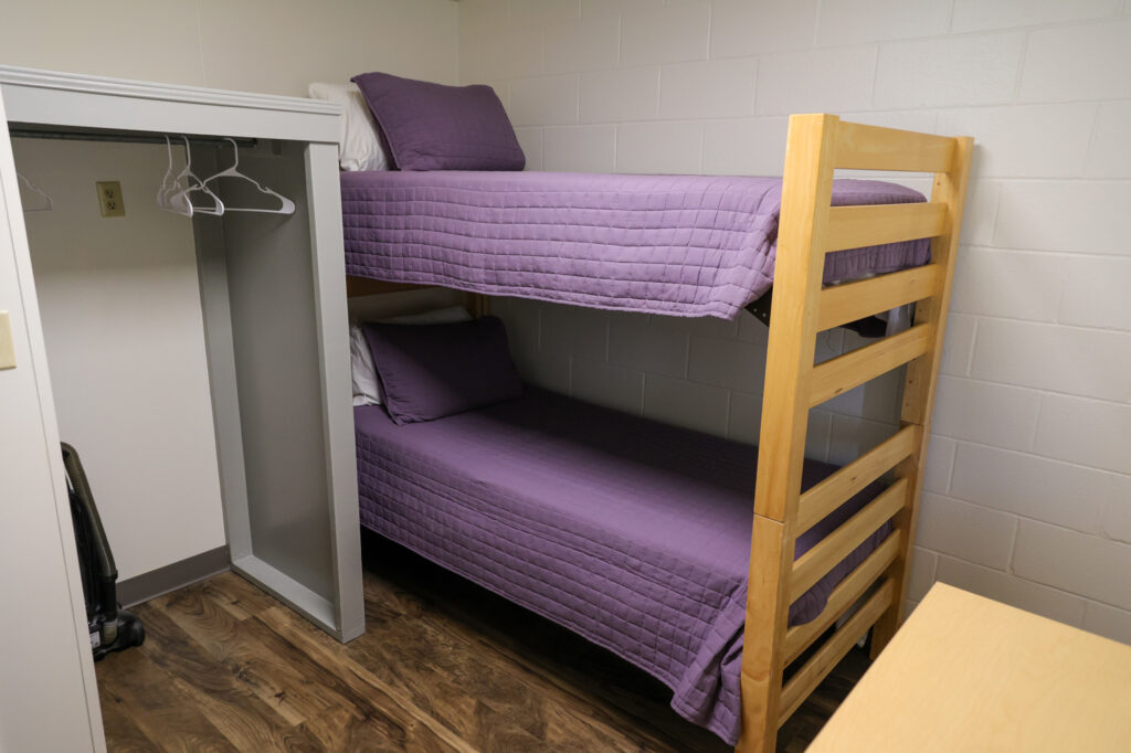 A small room with wooden bunk beds, both made with purple quilts and white pillows. An open closet with hangers and a vacuum is next to the beds. The room has white walls and a wood-patterned floor.