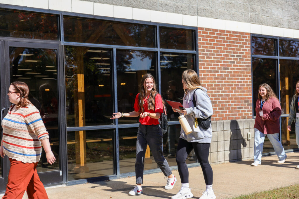 A group of students walk on a sidewalk next to a brick and glass building. Some carry bags, water bottles, or folders. Sunlight reflects off the windows. Trees and more students are visible in the background.