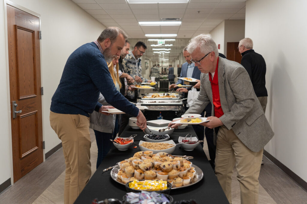 Several people serve themselves food from a buffet table in a hallway; the table holds pastries, fruit, and other dishes. The setting appears to be an indoor event or gathering with a business-casual dress code.
