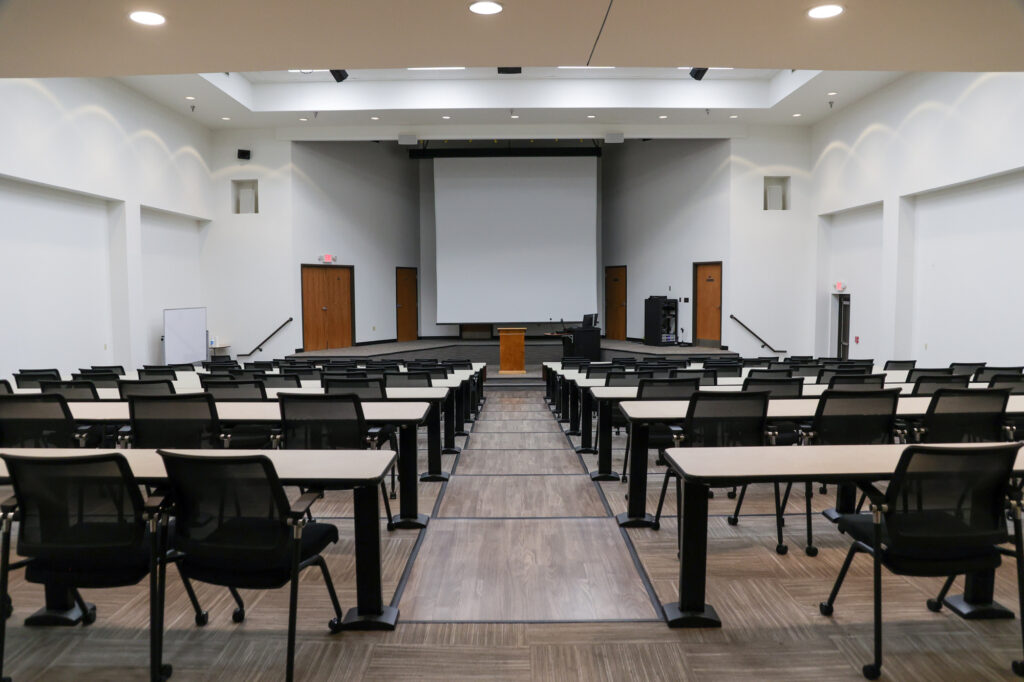 An empty lecture hall with rows of black chairs and long desks facing a wooden podium and large projection screen at the front. The room has white walls, brown doors, and a whiteboard on the left.