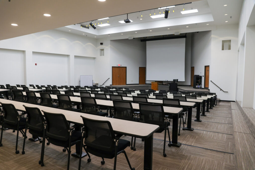 A modern lecture hall with rows of empty desks and black chairs facing a large projection screen and podium at the front. The room is well-lit with overhead lights and neutral colored walls and carpet.