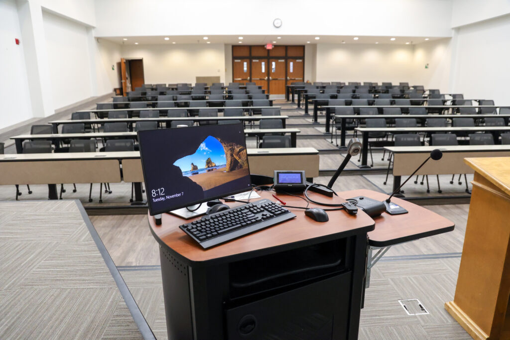 A lecture hall with rows of empty desks and chairs is shown from behind a podium. On the podium are a monitor, keyboard, mouse, microphone, and other electronic equipment. The room is well-lit and modern.