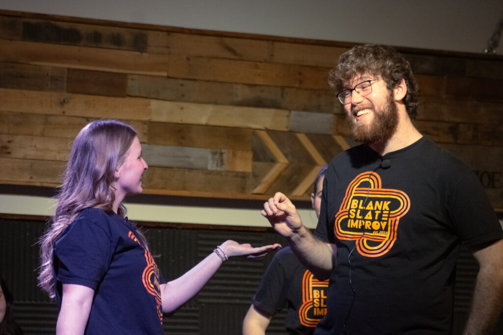 Two people wearing matching “Blank Slate Improv” shirts stand facing each other indoors. The woman is holding her hand out, and the man is smiling, gesturing with one hand. Both appear to be mid-conversation or performing.