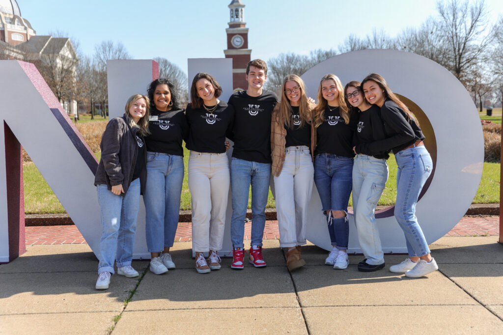 Eight young adults stand together in front of large campus letters, smiling and posing for a group photo outdoors. They wear matching black T-shirts and casual jeans, with a clock tower and trees visible in the background.