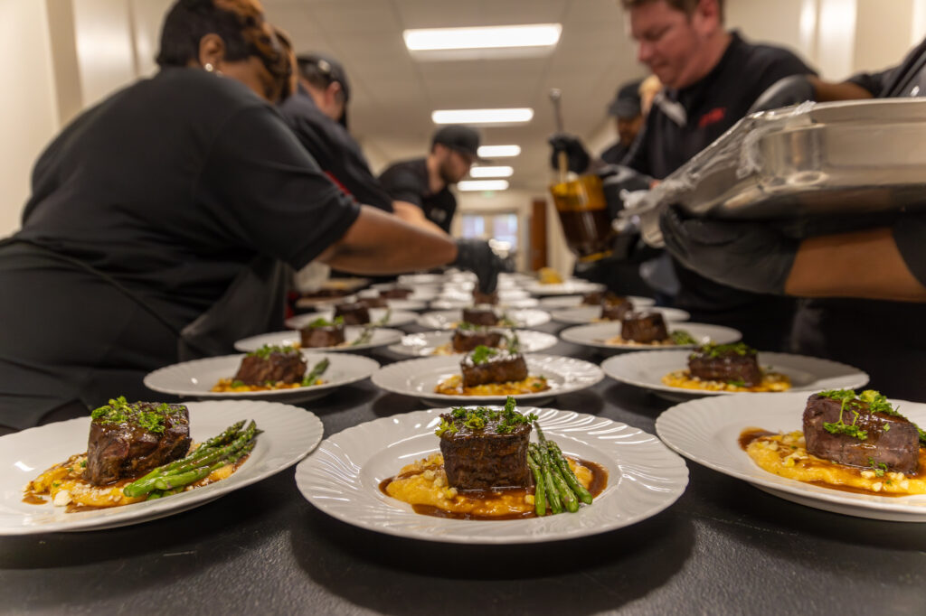 Chefs prepare and plate multiple servings of beef short ribs with asparagus and risotto in a professional kitchen, with dishes arranged in neat rows on a long table.
