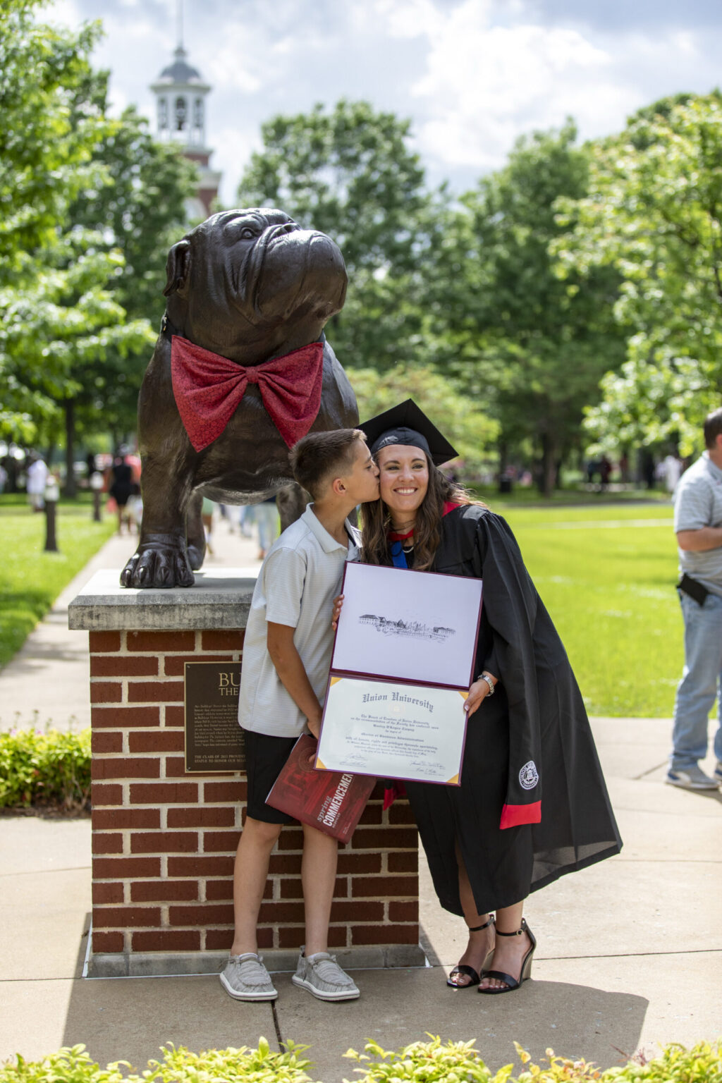 A woman in a graduation gown holds a diploma and smiles while a young boy kisses her cheek. They stand next to a large bulldog statue on a brick base outdoors, surrounded by cheering First-Year students, with people and trees in the background.