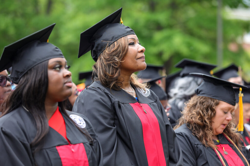 A group of graduates in caps and gowns stand outdoors during the Adult Studies ceremony. They wear black robes with red accents and tassels, and appear attentive. Trees and greenery are visible in the background, highlighting a proud moment following the admissions journey.