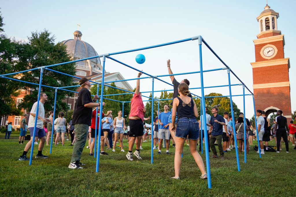 A group of people play a game with a blue ball inside a metal frame structure on a grassy field. Others watch or wait nearby. Buildings with domes and a clock tower are visible in the background.