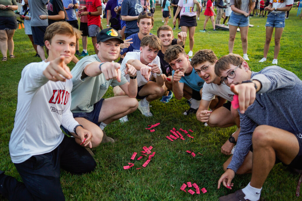 A group of young men kneel on grass, pointing toward the camera. They are surrounded by strips of red paper on the ground, and more people can be seen standing in the background at an outdoor event.