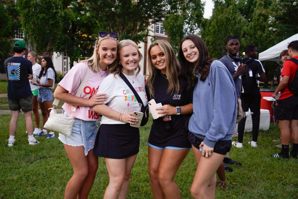 Four young women stand closely together, smiling at an outdoor event on a grassy area. People and a white tent are visible in the background. The women wear casual summer clothes and hold drinks. Trees and buildings surround the area.