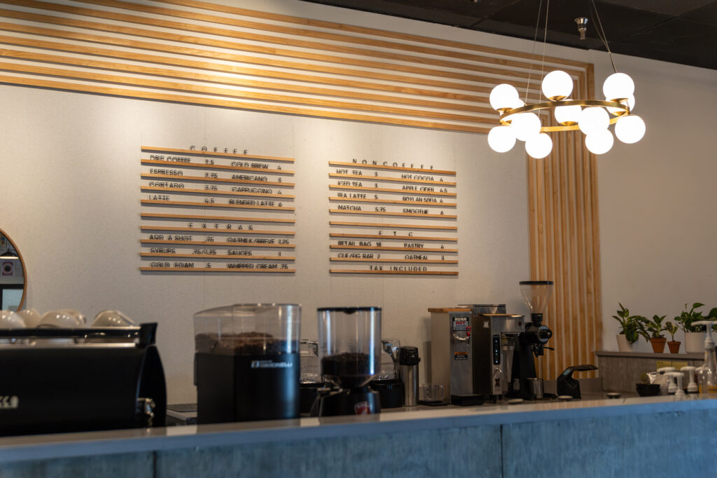 A modern coffee shop interior with a menu board listing coffee and non-coffee drinks, a coffee grinder, espresso machine, several plants, and a chandelier overhead. The counter is clean and tidy.