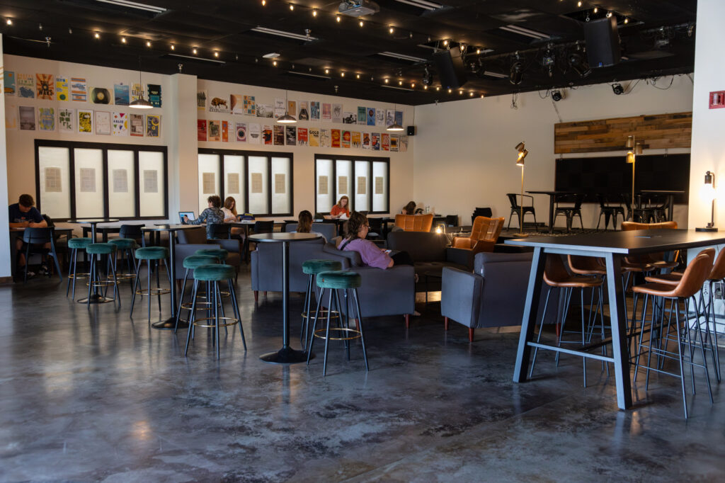 Spacious modern café with high ceilings, string lights, and people sitting at tables and sofas. Artworks line the upper walls, and a small stage with instruments is visible in the back. Large windows let in natural light.