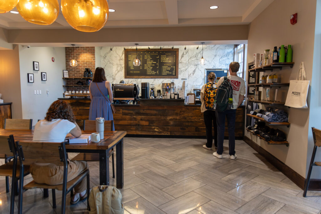 Three people stand at a coffee shop counter while one person sits at a table writing. Shelves with mugs and bottles are on the right wall. Three round ceiling lights hang above the wooden counter.