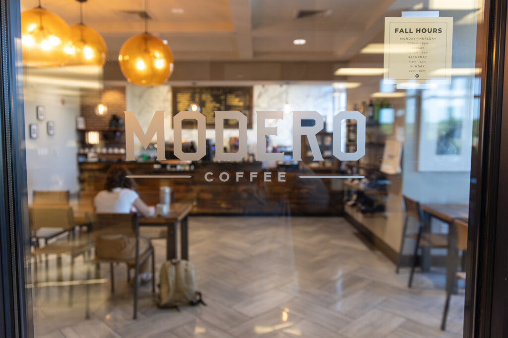 Glass door with Modero Coffee logo, showing a coffee shop interior. One person sits at a table with a laptop and backpack. Warm lighting, wooden furniture, and a sign with fall hours are visible.