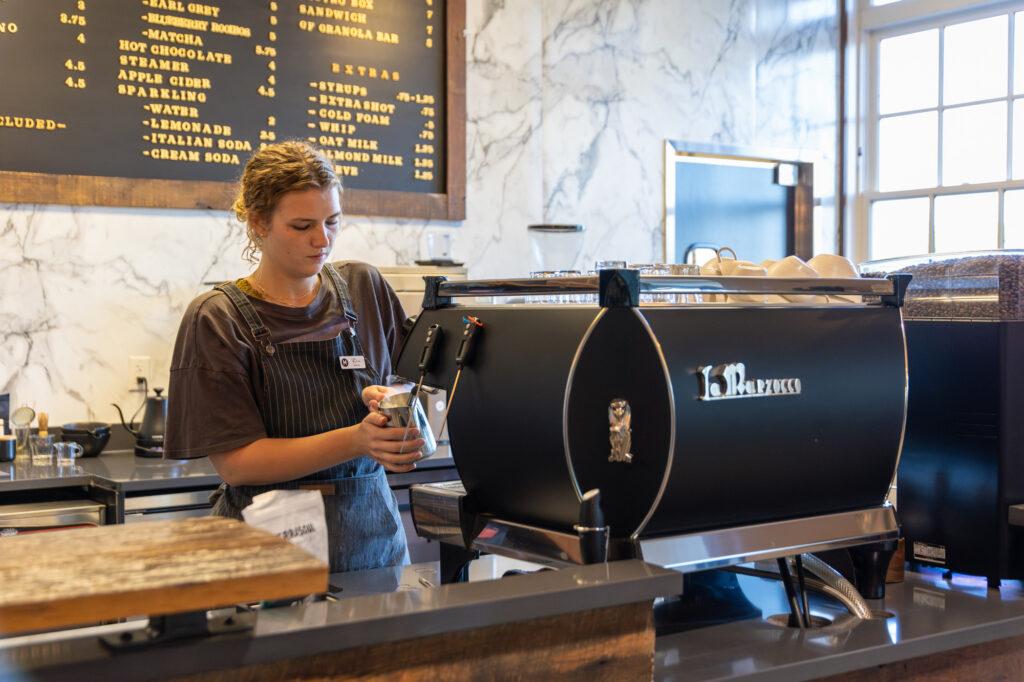 A barista wearing an apron steams milk using an espresso machine in a modern coffee shop. A menu board with drink options is visible on the wall behind her.