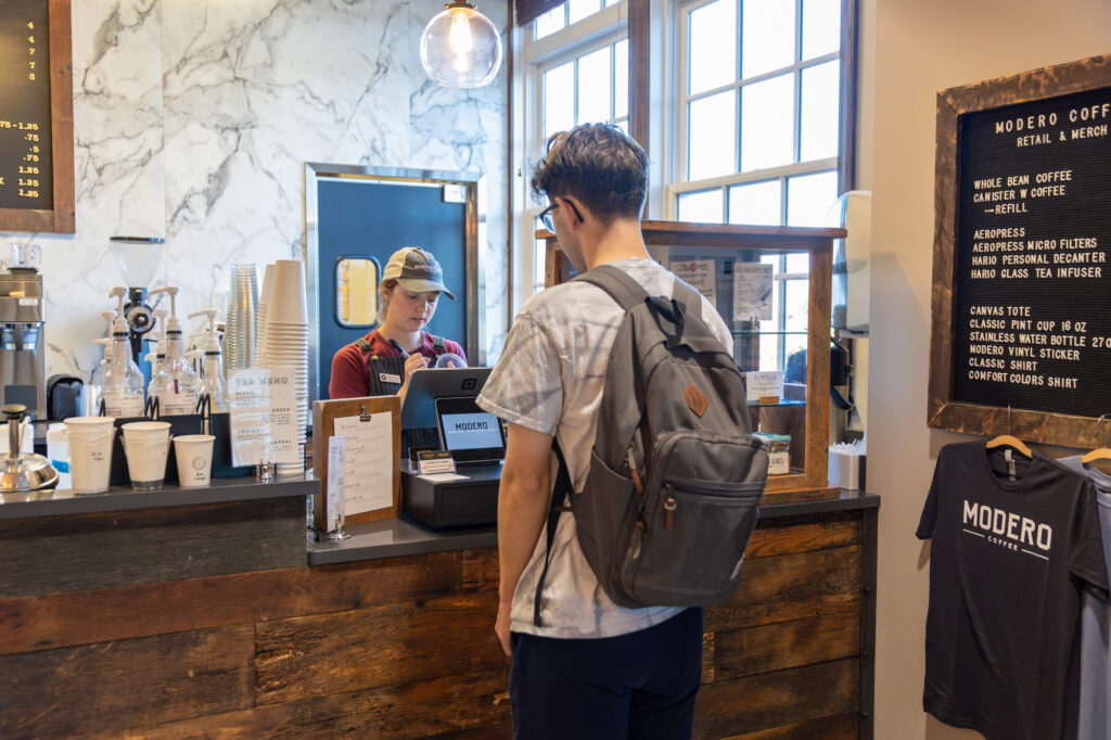 A person with a gray backpack stands at a wooden coffee shop counter, facing a barista using a tablet. The shop has menu boards, stacked cups, and a t-shirt display on the wall. Natural light enters through large windows.
