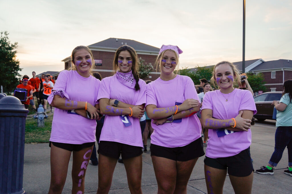 Four young women stand side by side outdoors, each wearing matching light purple shirts and black shorts, with words painted on their arms. Other people and buildings are visible in the background at dusk.