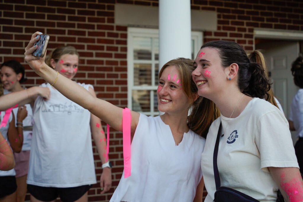 Two young women with neon face and arm paint smile and pose for a selfie outside a brick building. Other people with similar paint are in the background, engaging in conversation and activities.