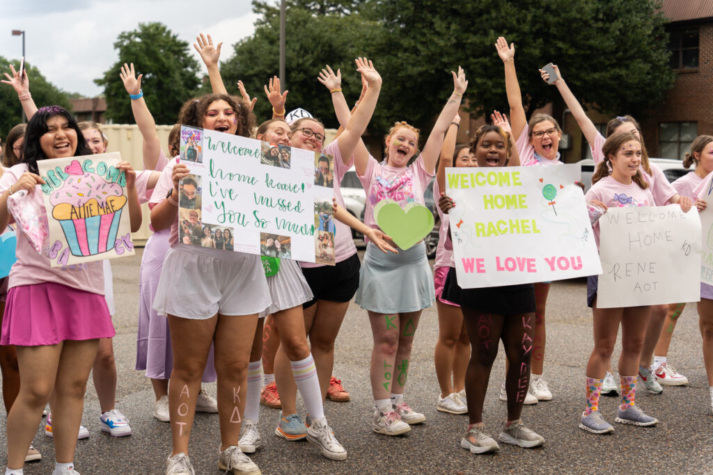 A group of young women outdoors hold welcome signs and raise their arms, smiling. Some wear pink and white outfits with Greek letters. Signs include messages like “Welcome Home Rachel” and “Welcome Home Rene.”.
