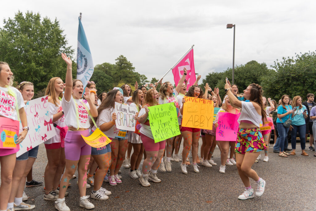 A group of young women, some holding colorful signs, cheer and raise their arms outdoors on a cloudy day. One woman stands facing them, leading the group. Trees and a few onlookers are visible in the background.