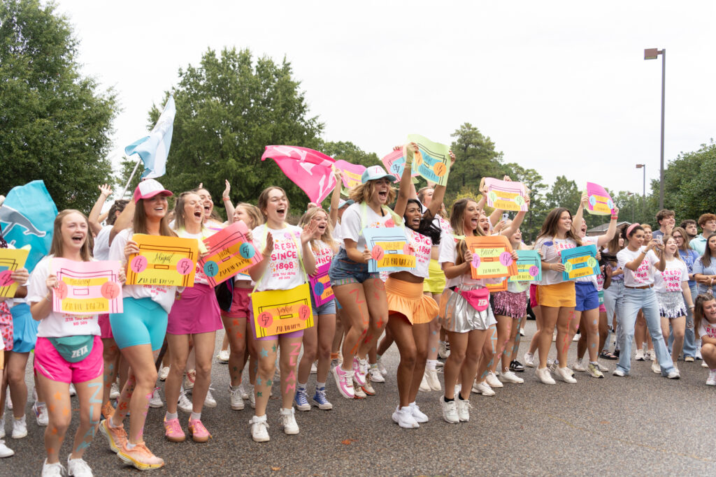 A group of young women stand outdoors, holding colorful signs and wearing matching T-shirts, shorts, and sneakers. Some are jumping or cheering, while others smile, with trees and people visible in the background.