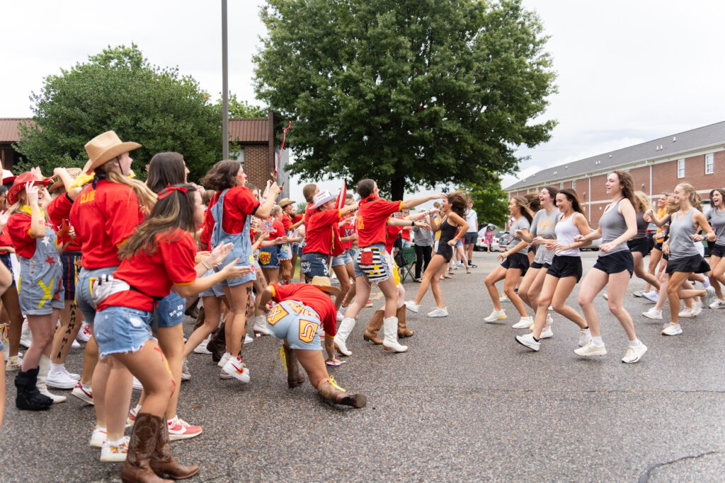 A large group of college-age people, some in red shirts and others in white shirts, energetically rush toward each other outside on a wet street near brick buildings and trees.