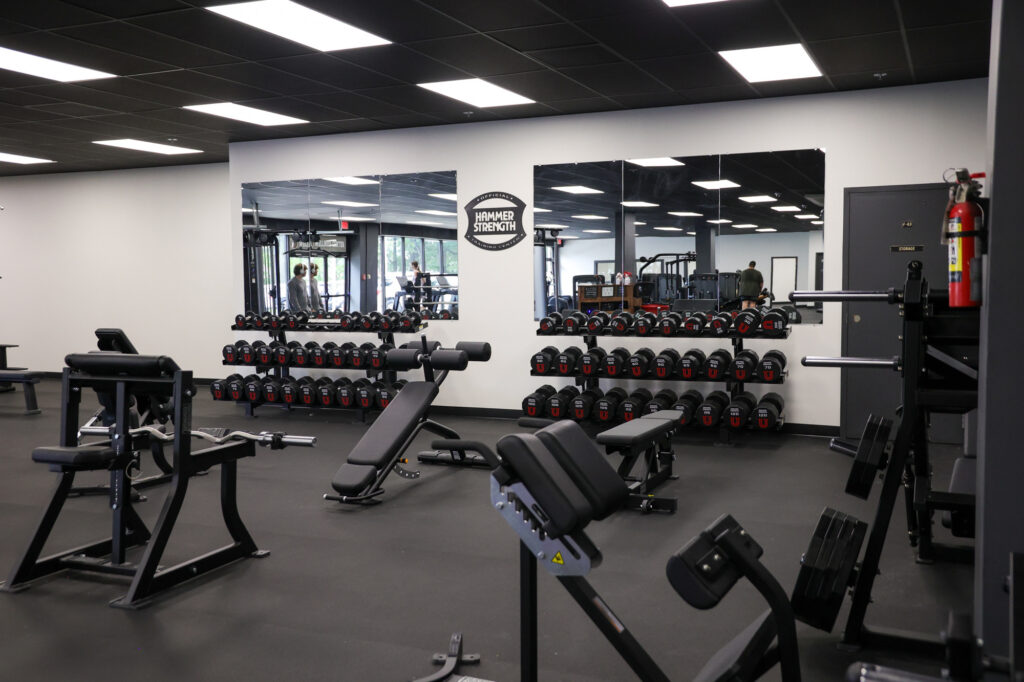 A gym with weight benches, racks of dumbbells, mirrored walls, and various strength training equipment. A fire extinguisher is mounted on the wall near a closed gray door. The floor is rubberized.