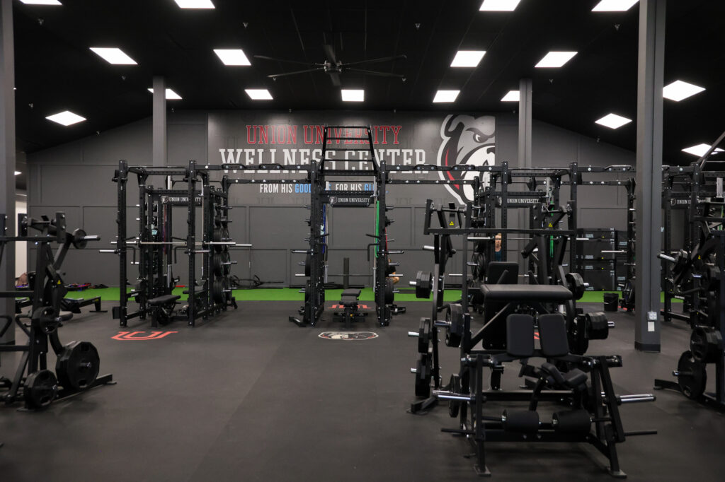 A fitness center with weightlifting equipment, squat racks, and benches. Union University Wellness Center is written on the back wall. The room is well-lit with black flooring and artificial turf along the walls.