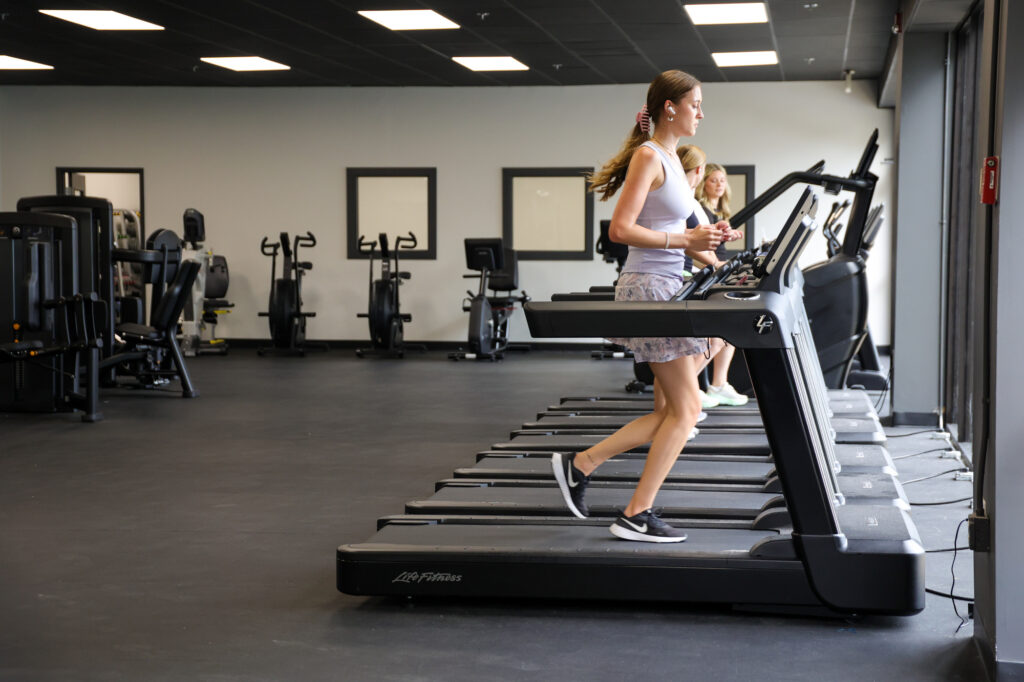 Two women are running on treadmills in a spacious gym with exercise bikes and weight machines in the background. The gym has large windows and a modern, minimalistic design.