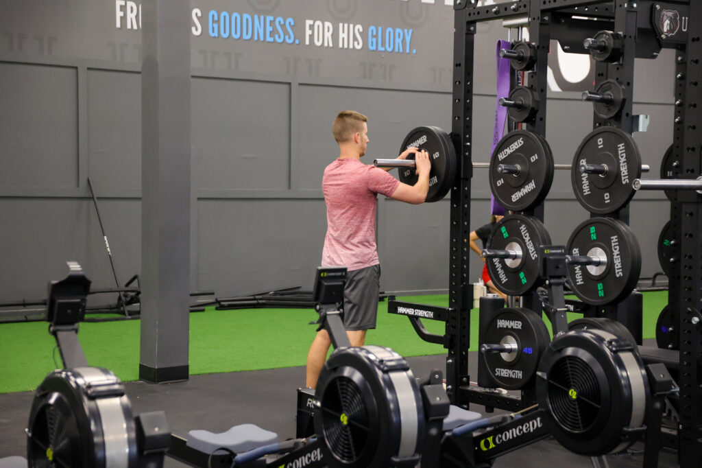 A person in a red shirt loads weights onto a barbell in a gym. Rowing machines and turf are visible, with motivational text on the wall in the background.