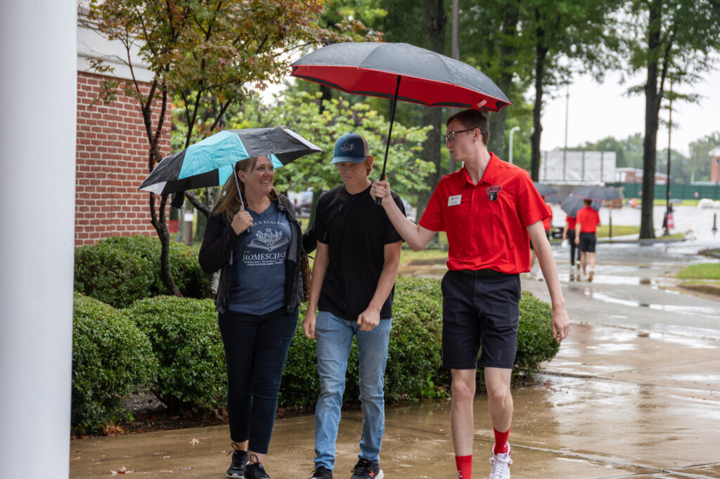Three people walk on a wet sidewalk under umbrellas; two share a black and teal umbrella, while the third holds a red one. A brick building, bushes, and trees are in the background.
