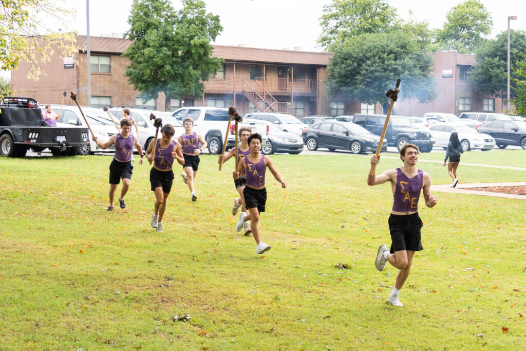 A group of young men in matching purple tank tops and black shorts run across a grassy area holding torches, with parked cars and a brick building in the background.