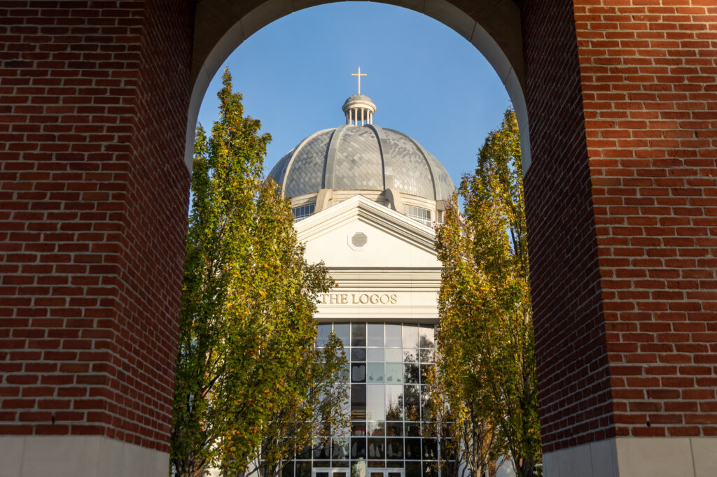 A domed building with a cross on top is framed by an archway and trees with green and yellow leaves. The building has large windows and the words VERITATE LOGOS on the front.