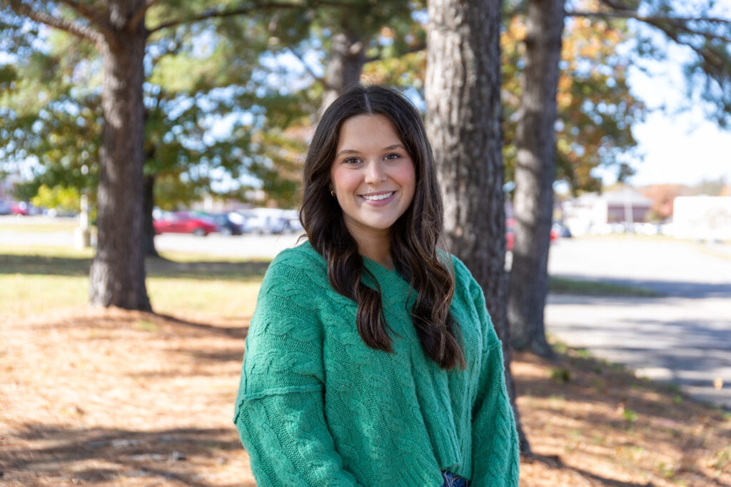 A young woman with long brown hair, wearing a green sweater, stands outdoors on a sunny day with trees and parked cars in the background. Smiling and facing the camera, Maggie Miller appears cheerful and relaxed in this vibrant scene.